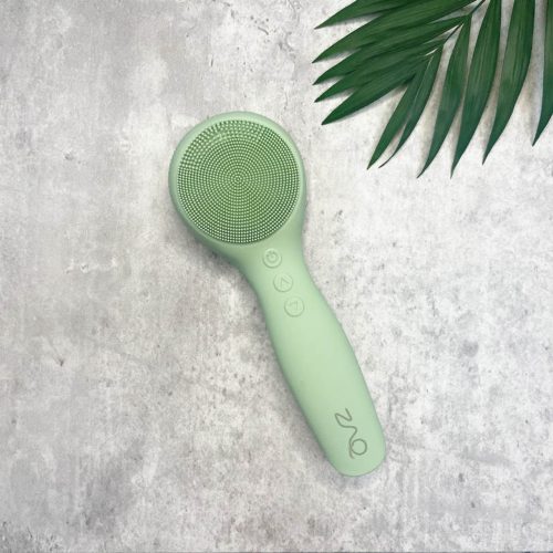 A mint green facial cleansing brush, one of the essential skincare tools, lying on a grey surface, accompanied by a green palm leaf at the top right corner.